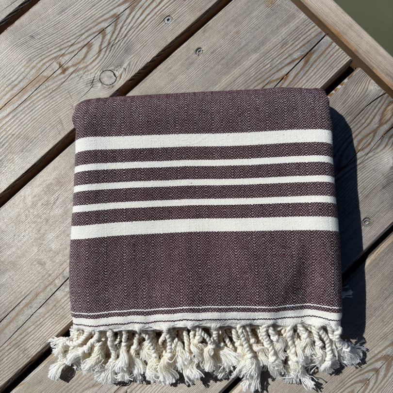 Hamamtuch ALMA – Dein vielseitiges Strandtuch für Sommer in Dunkelbraun zusammengelegt am Strand -DENIO