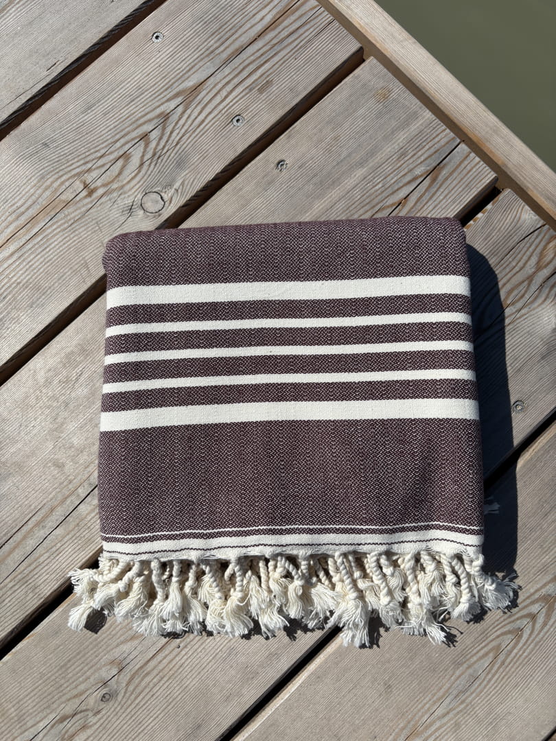 Hamamtuch ALMA – Dein vielseitiges Strandtuch für Sommer in Dunkelbraun zusammengelegt am Strand -DENIO