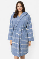 Frau trägt Bademantel in Farbe Saxblau aus glatter Baumwolle mit weißen Streifen und Kapuze vor weißem Hintergrund.